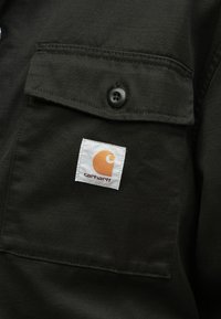 Ciemnozielona koszula z patką na kieszeni, ozdobiona czarnym guzikiem. Zawiera kwadratową białą etykietę z pomarańczowym logo i napisem "carhartt."