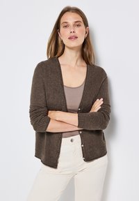 Brun, stickad cardigan med V-krage, ribbade ärmslut och knappstängning; bärs över en beige topp och matchas med ljusa byxor.