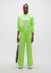 Γυναίκα με αθλητική ένδυση νέον πράσινου χρώματος κρατώντας ρακέτα τένις, στέκεται μπροστά σε λευκό απλό φόντο.