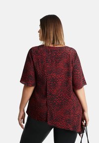 Blůza s leopardím potiskem v červené a černé barvě, krátké rukávy, volný střih, s asymetrickým lemem. Textura se zdá být hladká, z lehkého materiálu.