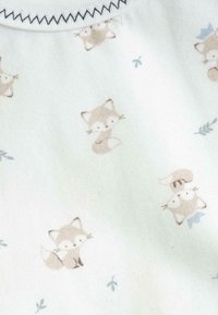 Tissu pastel clair présentant un motif répété de renards bruns et d'accents bleu clair. La texture est douce, avec un détail en bordure côtelée au niveau du col.