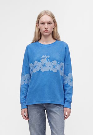 Jeune femme aux longs cheveux blonds portant un pull bleu avec de légers motifs floraux et le mot "ROXY" sur la poitrine.
