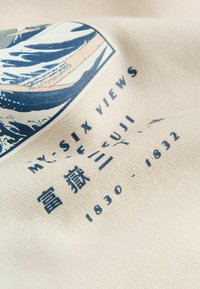 Nærbillede af stof med blå japansk inspireret bølgemønster, tekst "MY-SIX VIEWS," og datoer "1830 - 1832" på beige baggrund.