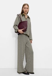 Blusa a scacchi bianco e nero e pantaloni a gamba larga, abbinati a una borsa tote intrecciata color bordeaux. Il completo presenta un dettaglio di collo verde.