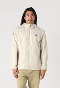 Beige kapuca jakna s prednjim zipom, elastičnim manžetami in stranskimi žepi. Ima majhen logotip na prsih in gladko, lahko tkanino.