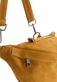 Gelbe Wildledertasche in dreieckiger Form, ausgestattet mit einem oberen Tragegriff, einem verstellbaren Riemen und silbernen Reißverschlussdetails. Weiche Textur mit sichtbaren Nähten.