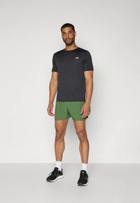 T-shirt atlética preta com um pequeno logótipo, combinada com calções verdes. Modelo usa ténis pretos e meias brancas. Design simples, estilo ajustado.