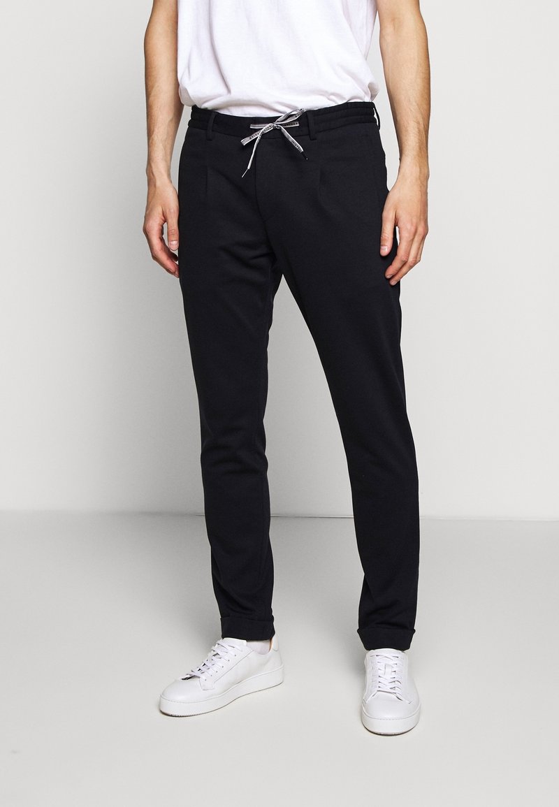 Zwarte slim-fit broek van zachte stof, met een elastische tailleband met trekkoord, gecombineerd met witte sneakers en een plain wit shirt.