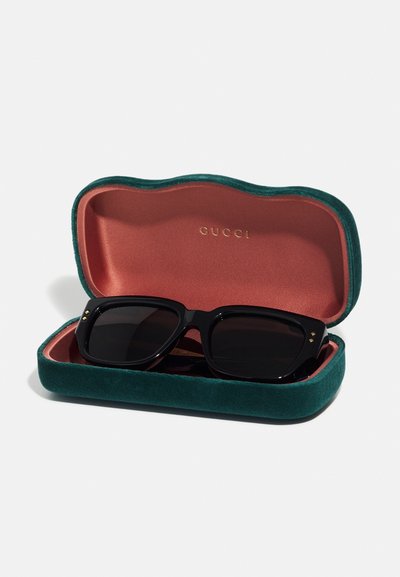 Black sunglasses in a green velvet case with a coral interior, featuring gold accents and a logo embossed inside the lid.