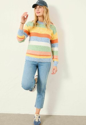 Kvinde i en stribet pasteltrøje, lyseblå jeans, blå kasket og sneakers poserer med det ene knæ løftet mod en almindelig hvid væg.