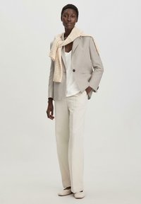 Valkoinen neulehuivi vaaleanharmaan bleiserin päällä, yhdistetty valkoiseen paitaan ja vaaleanbeigeihin leveisiin housuihin, luoden neutraalin, kerroksellisen asun.