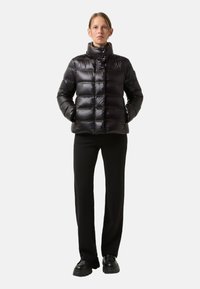 Schwarze Pufferjacke mit horizontalen Nähten, hohem Kragen und Druckknopfverschluss vorne. Das Modell trägt schwarze gerade geschnittene Hosen und klobige schwarze Schuhe.