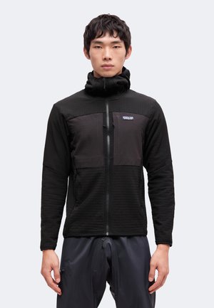 Jeune homme portant une veste noire zippée avec un col montant, debout face à l'avant, bras détendus le long du corps, sur un fond uni.