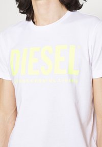 Bílý bavlněný tričko s kulatým výstřihem, zdobené velkým, živým žlutým nápisem "DIESEL" a "PRO ÚSPĚŠNÝ ŽIVOT" v tučném písmu.