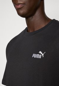 Sort bomulds T-shirt med rund halsudskæring og korte ærmer, med et hvidt broderet Puma-logo på brystet. Glat tekstur.