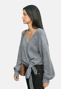 Suéter envolvente gris de punto con un profundo escote en V, mangas largas abullonadas y un detalle de cintura atada, fabricado en un material texturizado suave.