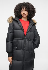 Zwarte pufferjas met een neopreen capuchon van nepbont, ritssluiting aan de voorkant en een trekkoord in de taille. Kenmerkt zich door een gestikte textuur en zijzakken. Rode binnenvoorkant van het sweatshirt zichtbaar.