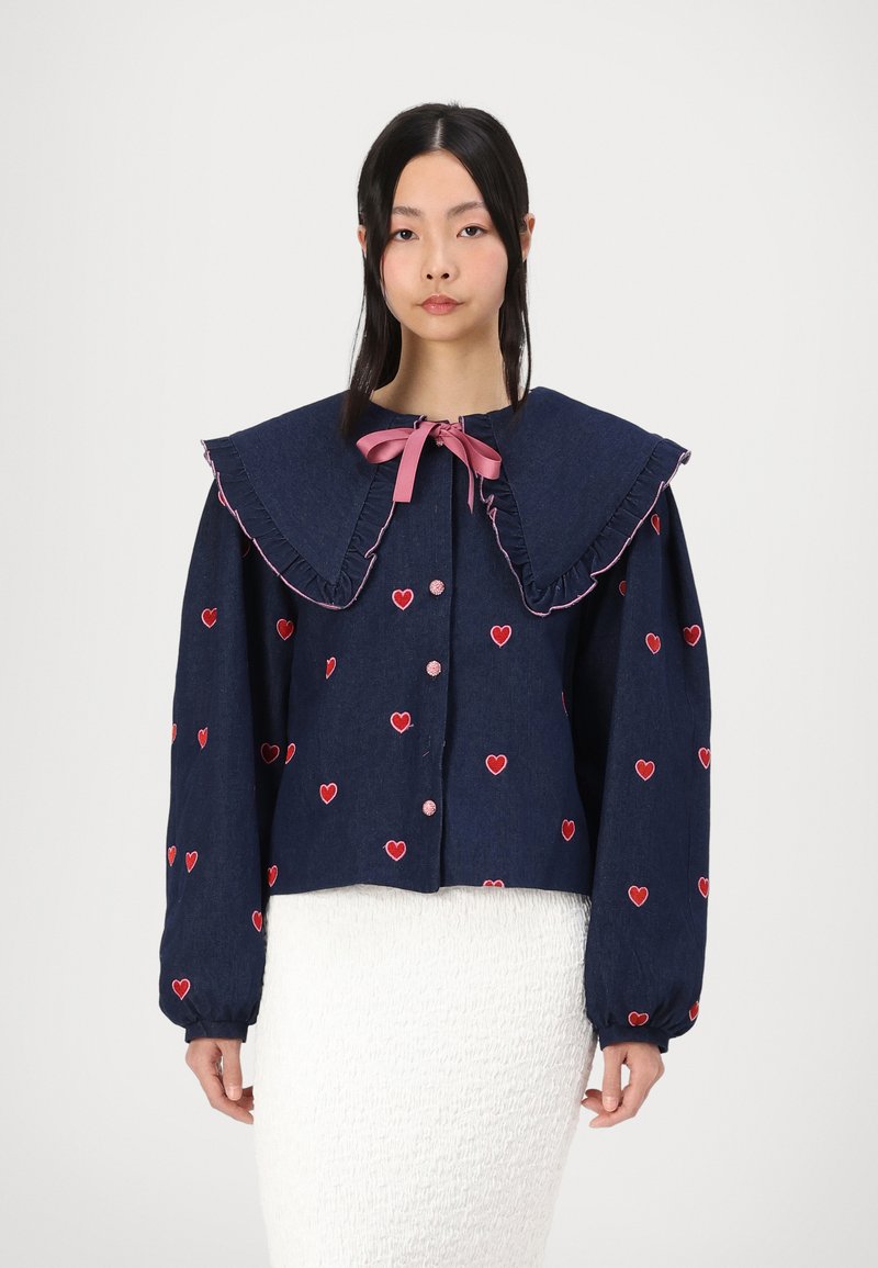 Ναυτικό τζάκετ από τζιν με μεγάλο φουσκωτό γιακά, ροζ κορδέλα και κεντημένη κόκκινη καρδιά. Έχει κλείσιμο με κουμπιά και φουσκωτά μανίκια.