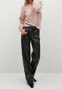 Frau trägt einen hellrosa Langarmpullover, der in hochgeschnittene schwarze Kunstlederweite Hosen gesteckt ist, und schwarze spitze Schuhe.