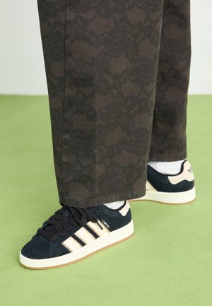 Baskets Adidas Campus noires avec des bandes blanches, portées avec des chaussettes blanches et un pantalon sombre à motifs sur un sol vert clair.