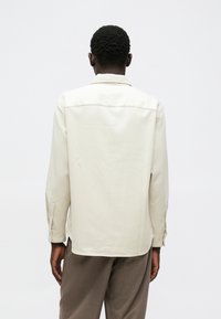 Chemise beige clair à manches longues, avec un col pointu et des accents de bouton. Présente une surface texturée et un ourlet arrondi.