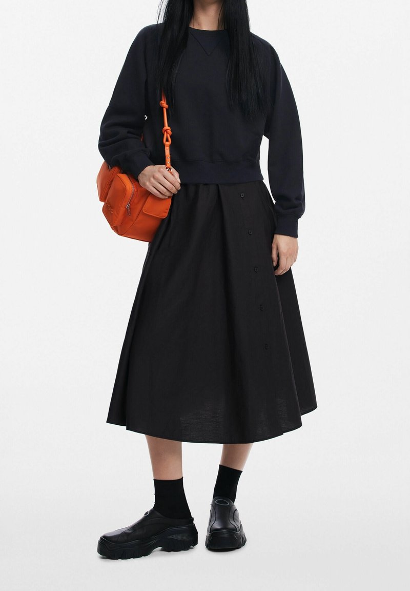 Femme portant un haut noir à manches longues et une jupe, des chaussettes noires, des chaussures à plateforme noires, tenant un sac à bandoulière orange avec une sangle nouée.