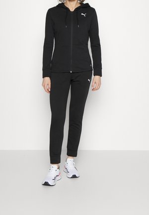 Femme portant une veste zippée à capuche noire Puma et un pantalon assorti avec des chaussures de sport blanches, debout devant un fond blanc.