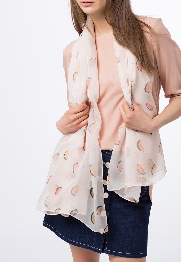 Femme portant une écharpe blanche transparente avec des motifs de fruits roses et verts, sur un haut rose pâle et une jupe en denim bleu foncé à boutons.