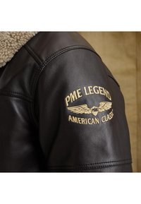 Čierna kožená bunda s ramenami a béžovým sherpa golierom, so zlatou výšivkou textu "PME Legend American Classic" a emblémom krídla.