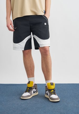 Persoon die zwarte en witte Nike-short draagt, een beige shirt, witte sokken en grijs-wit-gele hoge Nike-sneakers, staand op een blauw tapijt.