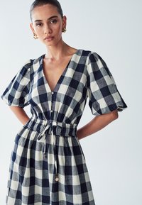 Vestido a cuadros con escote en V, mangas abullonadas, cintura elástica y cordón. Los colores incluyen azul marino y crema, fabricado con una tela ligera.