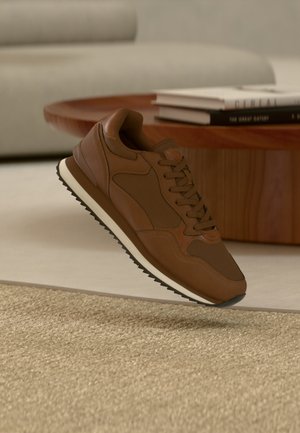 Zapatilla marrón con suela blanca suspendida sobre una alfombra beige delante de una mesa de madera con libros apilados y un sofá gris claro de fondo.