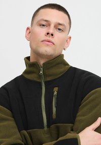 Fleece genser med svart og olivengrønn fargeblokkdesign. Har høy krage, glidelåslomme på brystet og teksturert stoff.