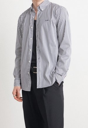 Person wearing a white and black vertically striped button-up shirt over a black top, with black pants and a black belt, hand in pocket.