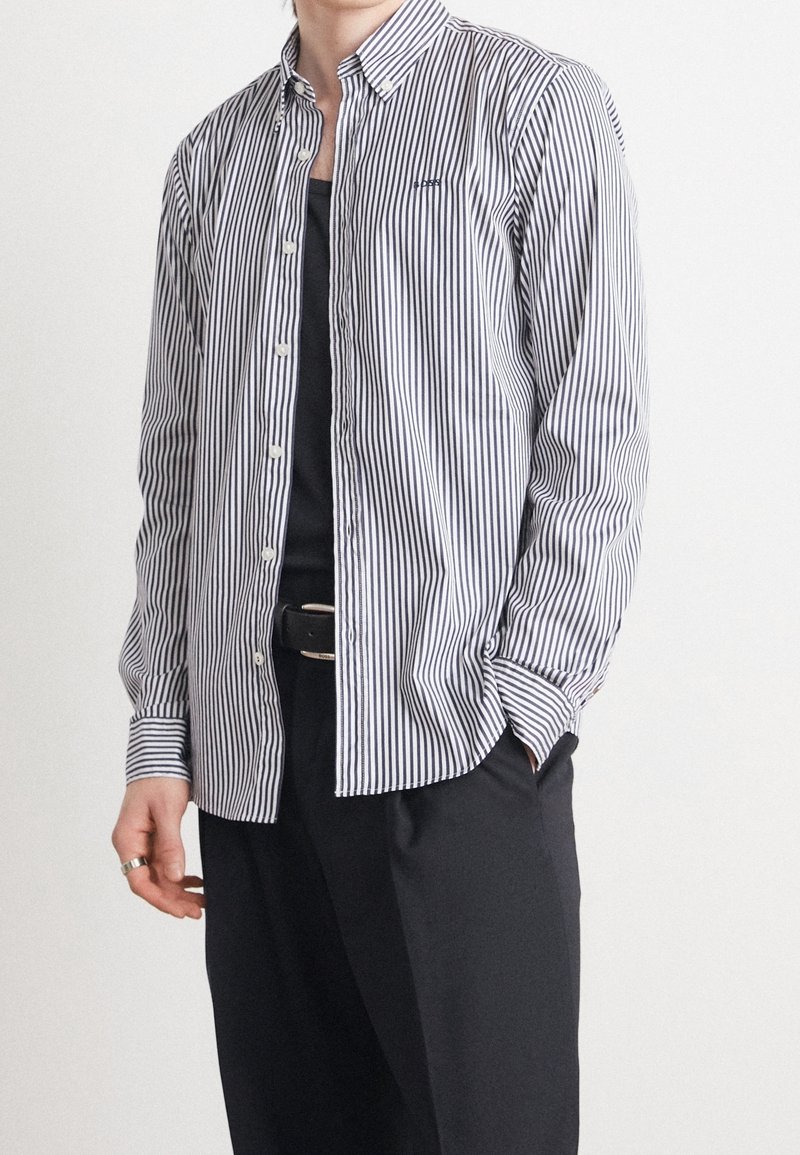 Personne portant une chemise boutonnée blanche et noire à rayures verticales par-dessus un haut noir, avec un pantalon noir et une ceinture noire, main dans la poche.