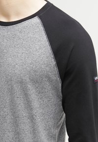 Närbild av en person som bär en grå och svart långärmad tröja med synlig sömnad och ett litet logomärke på den övre ärmen.