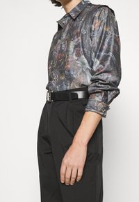 Bluse mit floralem Muster in dunklen Farbtönen, ausgestattet mit einem Kragen, Knopfverschluss und langen Ärmeln mit aufgerollten Manschetten. Kombiniert mit hochgezogenen schwarzen Hosen.