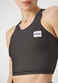 Top desportivo preto, com decote redondo, alças largas e um rótulo branco proeminente com o nome da marca na área do peito.