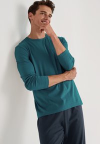 Teal lange mouwen shirt met een ronde hals en een borstzak, gecombineerd met donkere broek. Glad textuur, casual pasvorm en relaxte stijl.