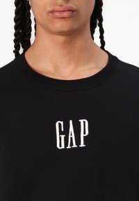 Fekete pamut kereknyakú pulóver, amelyen fehér hímzett "GAP" felirat található a mellrészen, puha tapintású és klasszikus szabású.
