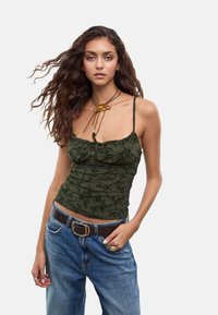 Jeune femme aux longs cheveux ondulés portant un haut à bretelles spaghetti floral vert foncé, un jean bleu avec une ceinture marron et un collier en fleurs.
