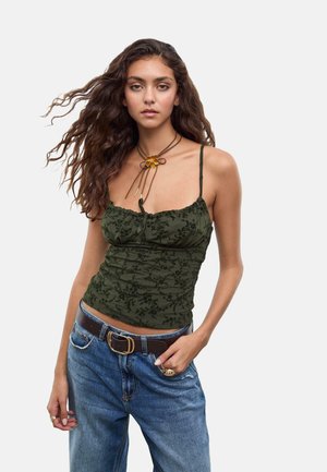Giovane donna con lunghi capelli ondulati, che indossa un top a spaghetto floreale verde scuro, jeans blu con una cintura marrone e una collana a forma di fiore.