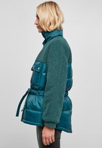Veste matelassée turquoise avec partie supérieure en polaire, poches à pression et taille ceinturée. Comprend un col haut et un tissu texturé sur les manches.