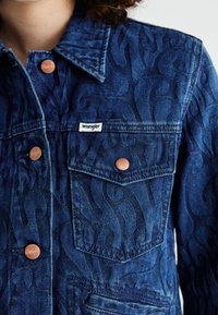 Chaqueta de mezclilla con patrón de remolinos en relieve, color azul profundo, botones en tono cobre y etiqueta de la marca en el bolsillo del pecho.