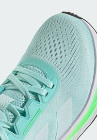 Zapatilla deportiva de color azul claro con parte superior de malla, cordones planos, detalles texturizados y un acento verde contrastante en la suela para mayor soporte.