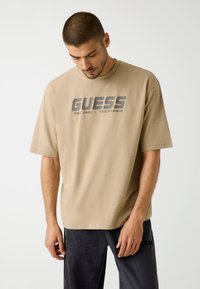 Beige cotton t-shirt with short sleeves, featuring a dark gray "GUESS" logo and "EST. 1981 - CALIFORNIA" text on the front.