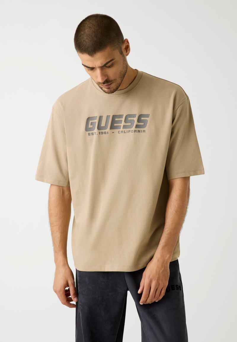 Beige cotton t-shirt with short sleeves, featuring a dark gray "GUESS" logo and "EST. 1981 - CALIFORNIA" text on the front.