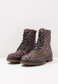 Par de botas marrones hasta el tobillo con estampado de leopardo negro, cordones negros, cremalleras laterales y suelas gruesas y translúcidas sobre una superficie blanca.