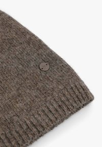 Suéter de lana marrón tejido, con dobladillo acanalado y una etiqueta redonda de tela marrón con un emblema de calavera. Textura suave y material cálido.