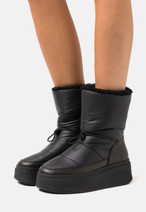 Bottes d'hiver noires isolées avec semelles épaisses compensées et cordon de serrage réglable à la cheville, portées sur des jambes nues sur fond blanc.